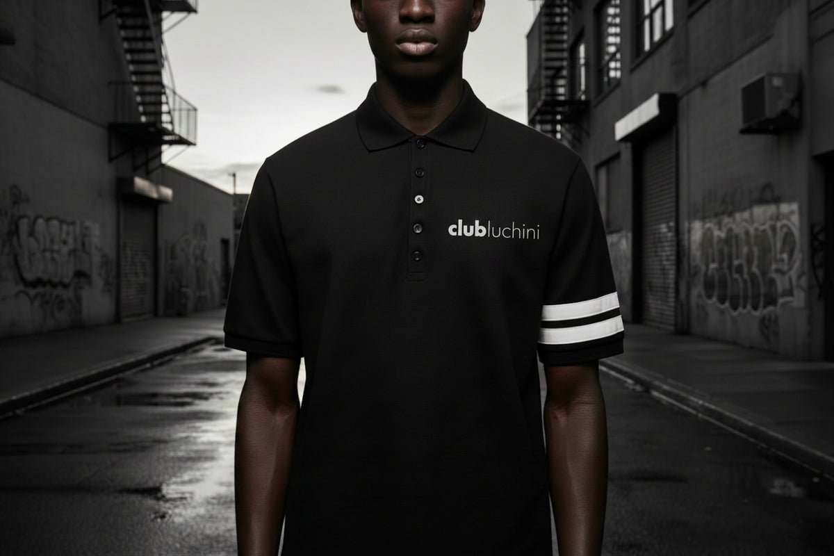 A male model wearing a black Club Clutchini polo shirt with white logo text on the chest and white stripes on the sleeve, standing in a wet urban alleyway.