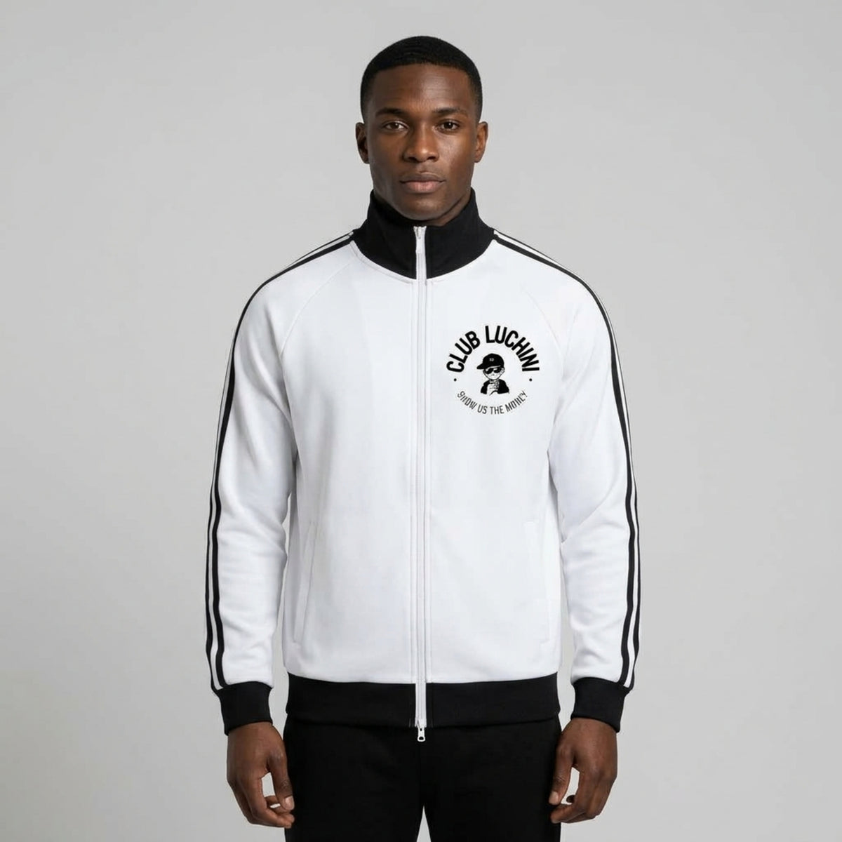 A man wearing a white Club Luchin track jacket with black trim, black ribbed cuffs and hem, and a black circular chest graphic, paired with black pants, against a light gray studio background.